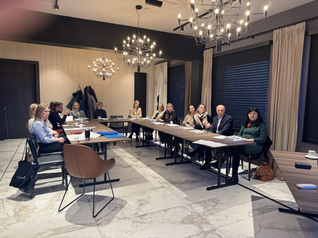 Grupa osób siedzi przy stole konferencyjnym w kształcie litry U.