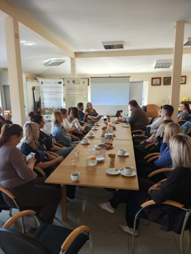 Grupa osób siedzi przy podłużnym stole po obu stronach. Wszyscy patrzą w stronę wyświetlonej na ekranie projekcyjnym prezentacji.