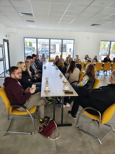 Duża grupa osób siedzi przy stołach połączonych w kształt litery L. Większość osób ma przed sobą filiżankę kawy.