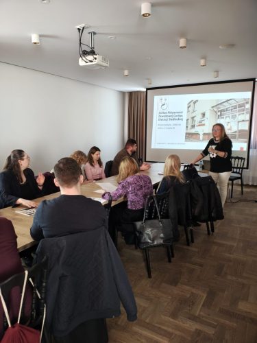 Grupa osób siedzi przy podłużnym stole po obu stronach. Wszyscy patrzą w stronę kobiety prowadzącej spotkanie oraz prezentacji która jest wyświetlona za nią.