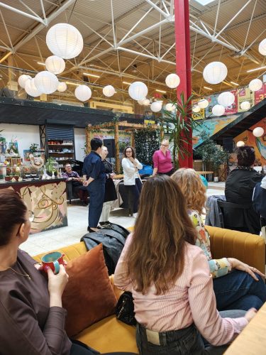 Grupa osób w dużej, otwartej przestrzeni przypominającej restauracje lub kawiarnie. Część osób siedzi na żółtej kanapie, część siedzi przy stole, kilka osób stoi obok baru.