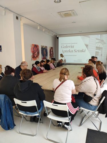 Grupa osób siedzi przy szerokim, podłużnym stole. Na przeciwko, na ekranie projekcyjnym jest wyświetlona prezentacja. Obok na ścianie wiszą 3 obrazy.