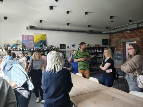 Grupa osób chodząca po pomieszczeniu przypominającym pracownie malarską lub garncarską. Przy ścianie stoją obrazy i ceramika.