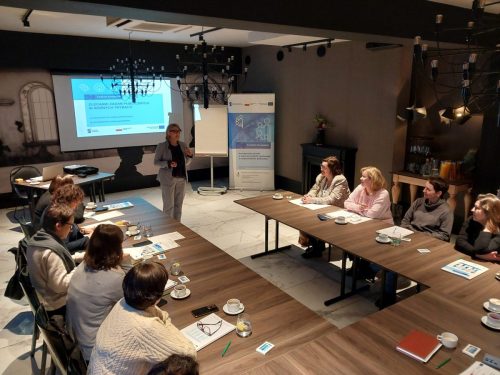 Grupa osób siedzi przy łączonych stołach w kształt litery U. Na środku sali stoi kobieta prowadząca szkolenie. W tle jest wyświetlona prezentacja.