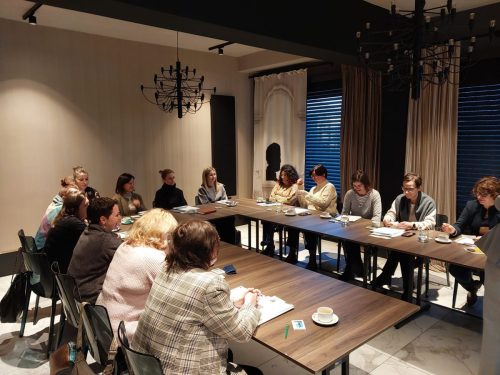 Grupa osób siedzi przy łączonych stołach w kształt litery U.