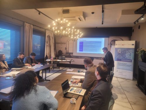 Grupa osób siedzi przy łączonych stołach i patrzy w kierunku wyświetlonej prezentacji. Obok stoi mężczyzna prowadzący szkolenie.