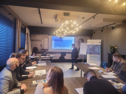 Grupa osób siedzi przy łączonych stołach i patrzy w kierunku wyświetlonej prezentacji. Obok stoi mężczyzna prowadzący szkolenie. Kadr 2.