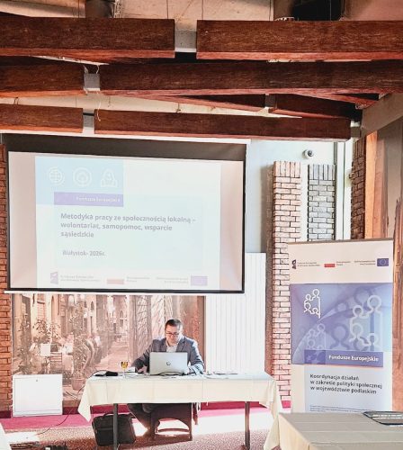 Prowadzący szkolenie siedzi przy stole z laptopem. Nad nim jest wyświetlona prezentacja.