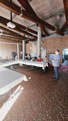 Grupa osób siedzi przy łączonych stołach w trakcie szkolenia. Przed nimi stoi prowadzący.