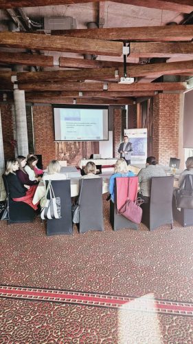 grupa osób siedzi przy łączonych stołach w trakcie szkolenia. W tle jest wyświetlona prezentacja.