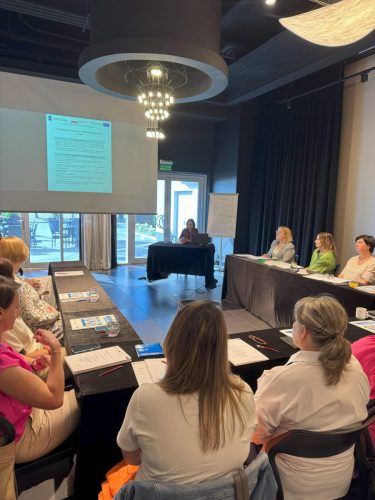 Sala z stołami ustawionym w kształt litery U przy których siedzą uczestnicy szkolenia. Na drugim planie widać ekran projekcyjny z wyświetloną prezentacją obok stoi stół przy którym siedzi osoba prowadząca szkolenie.
