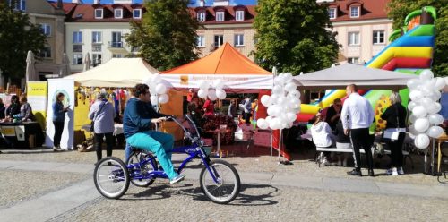 Stoiska podmiotów ekonomii społecznej wystawione w jednym rzędzie na rynku Kościuszki w Białymstoku z okazji Targów Ekonomii Społecznej. Przed stoiskami przejeżdża mężczyzna na rowerze trójkołowym.