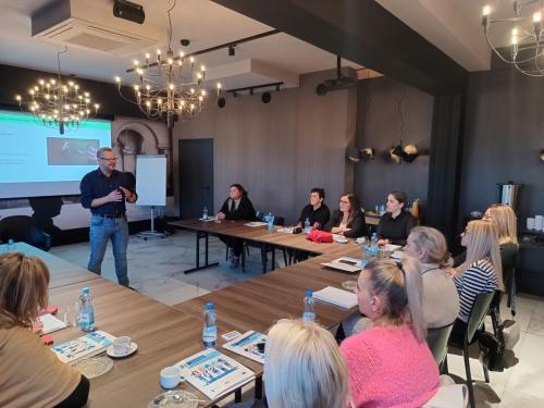 Grupa ludzi siedzących przy stole konferencyjnym, słuchających osoby prowadzącej szkolenie. W tle za prowadzącym wyświetlana jest prezentacja na ekranie projekcyjnym. Stół jest zastawiony butelkami z wodą, kubkami i dokumentami.