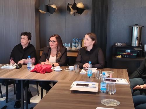 Trzy kobiety siedzą obok siebie przy stole konferencyjnym. Wszystkie trzy kierują wzrok w lewą stronę na osobę prowadzącą szkolenie. Na stole stoją butelki wody, filiżanki oraz dokumenty.