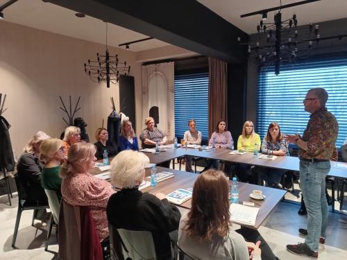 Uczestników siedzących wokół stołu konferencyjnego. Na środku stoi osoba prowadząca prezentację. Na stole widoczne są butelki z wodą, kubki, notatki i dokumenty.