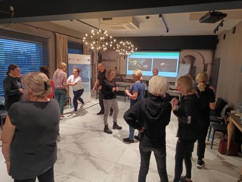 Grupa ludzi w pomieszczeniu, które biorą udział w parach w fizycznej aktywności. Na przedniej ścianie pokoju znajduje się ekran projekcyjny, a także widoczny jest baner z tekstem i logotypami.