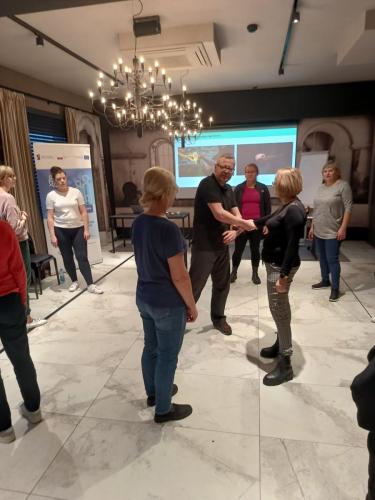 Grupa ludzi w pomieszczeniu z dużym ekranem wyświetlającym prezentację. Uczestnicy stoją rozproszeni na sali, a na środku stoi prowadzący stoi prowadzący który pokazuje chwyt na jednej z uczestniczek.