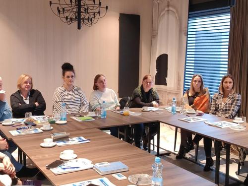Grupa osób siedzi przy stole konferencyjnym ustawionym w kształt litery U i patrzy w jednym kierunku.