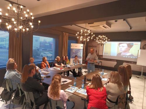 Grupa uczestników siedzi przy stole konferencyjnym i słucha prowadzącego który stoi na środku i przemawia. W tle wyświetlone nagranie wideo na ekranie projekcyjnym.