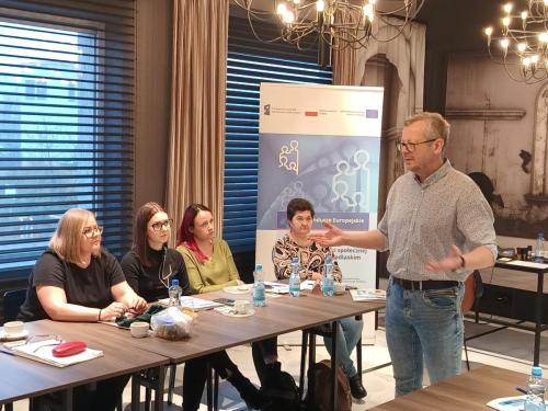 Cztery kobiety siedzą przy stole i patrzą na prowadzącego który przemawia do reszty grupy. W tle baner projektu.