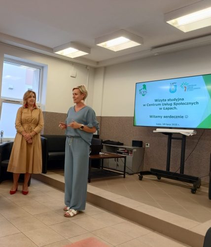 Dwie kobiety na tle prezentacji wyświetlonej na telewizorze. Jedna z kobiet przemawia.