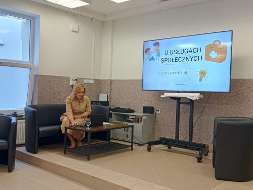 Sala z telewizorem na którym jest wyświetlona prezentacja. Obok na czarnej kanapie siedzi kobieta robiąca coś na laptopie.