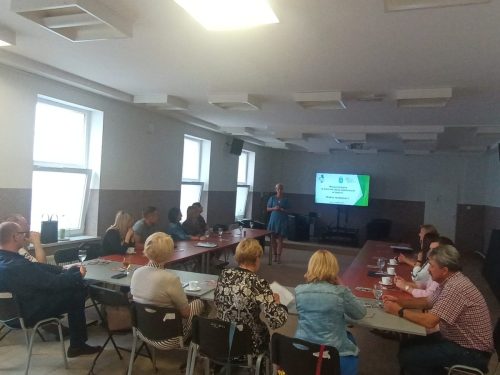Sala z łączonymi stołami w kształt litery U przy których siedzą uczestnicy. Na środku stoi kobieta prowadząca a za nią stoi telewizor z wyświetloną prezentacją.