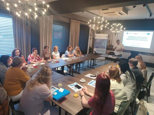 Sala z łączonymi stołami w kształt litery U przy których siedzą uczestnicy spotkania. Na środku sali stoi mężczyzna który przemawia. W tle jest wyświetlona prezentacja.