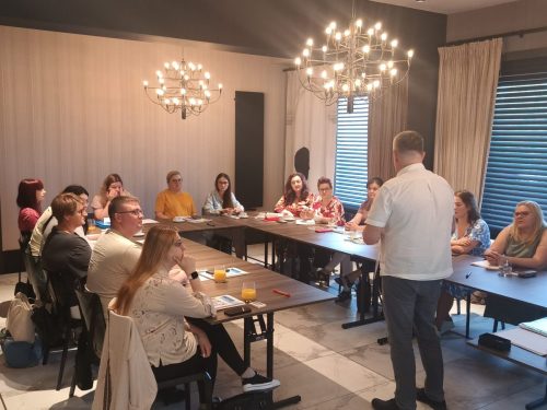Sala z łączonymi stołami w kształt litery U przy których siedzą uczestnicy spotkania. Na środku sali stoi mężczyzna.