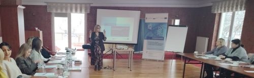 Kobieta która prowadzi szkolenie przemawia do uczestników spotkania którzy siedzą po bokach sali. W tle wyświetlona prezentacja.