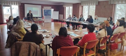 Grupa osób siedzi przy łączonych stołach i patrzą w kierunku kobiety która prowadzi szkolenie. Kadr 2.