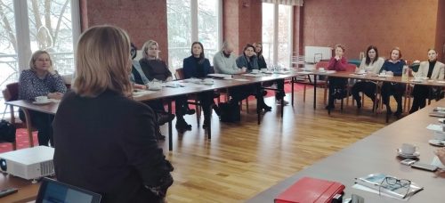 Grupa osób siedzi przy łączonych stołach i patrzą w kierunku kobiety która prowadzi szkolenie.