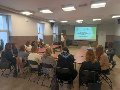 Grupa osób siedzi przy łączonych stołach w trakcie szkolenia. Na końcu sali wisi ekran projekcyjny z wyświetloną prezentacją.