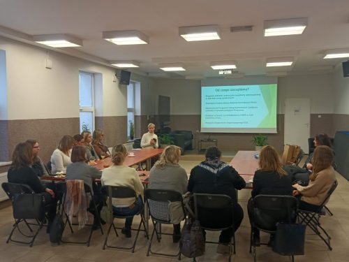 Grupa osób siedzi przy łączonych stołach w trakcie szkolenia. Na końcu sali wisi ekran projekcyjny z wyświetloną prezentacją (kadr 2).