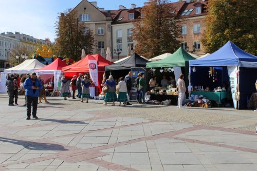 Stoiska podmiotów ekonomii społecznej wystawione w jednym rzędzie na rynku Kościuszki w Białymstoku z okazji Targów Ekonomii Społecznej. Przy namiotach chodzą ludzie.