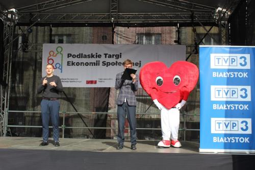 scena targów ekonomii społecznej na niej prezenter tłumacz migowy maskotka targów oraz patronat medialny telewizja regionalna oddział w białymstoku