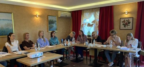 Sala z łączonymi stołami w kształt litery L przy których siedzą uczestnicy szkolenia. Na środku stoi mały stolik z projektorem.