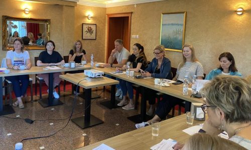 Sala z stołami ustawionymi w kształt litery U przy których siedzą uczestnicy spotkania. Na środku stoi mały stolik z projektorem.