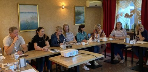 Sala z stołami ustawionymi w kształt litery U przy których siedzą uczestnicy spotkania. Na środku stoi mały stolik z projektorem.