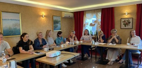 Sala z łączonymi stołami w kształt litery L przy których siedzą uczestnicy szkolenia. Na środku stoi mały stolik z projektorem.