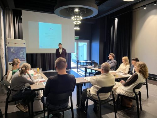 Grupa osób siedzi przy łączonych stołach w trakcie szkolenia. Na środku na tle wyświetlonej prezentacji mężczyzna prowadzący szkolenie.