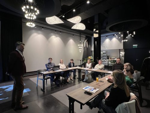 Grupa osób siedzi przy łączonych stołach w trakcie szkolenia. Przed uczestnikami stoi mężczyzna prowadzący szkolenie.