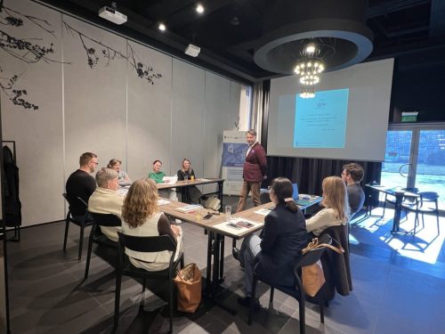 Grupa osób siedzi przy łączonych stołach w trakcie szkolenia. Przed uczestnikami stoi mężczyzna prowadzący szkolenie. W tle jest wyświetlona prezentacja.