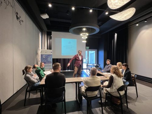 Grupa osób siedzi przy łączonych stołach w trakcie szkolenia. Na środku na tle wyświetlonej prezentacji mężczyzna prowadzący szkolenie który przemawia do uczestników.