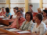 Publiczność siedzi w ławkach w kilku rzędach. Na ławkach leża teczki z dokumentami i długopisy.