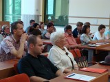 Publiczność siedzi w ławkach i patrzy w jednym kierunku. Na ławkach leżą teczki i dokumenty.