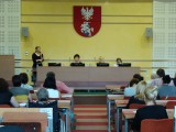 Sala wykładowa z herbem województwa podlaskiego. Pod herbem znajdują się cztery osoby prowadzące, jedna z nich stoi i przemawia do publiczności siedzącej na przeciwko w ławkach.