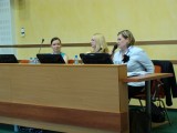 Trzy kobiety siedzą przy stole z ekranami i mikrofonami. Jedna z nich przemawia przez mikrofon.