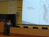 Kobieta stoi przy mównicy i przemawia do mikrofonu. Obok jest wyświetlana prezentacja o budowie kręgosłupa.
