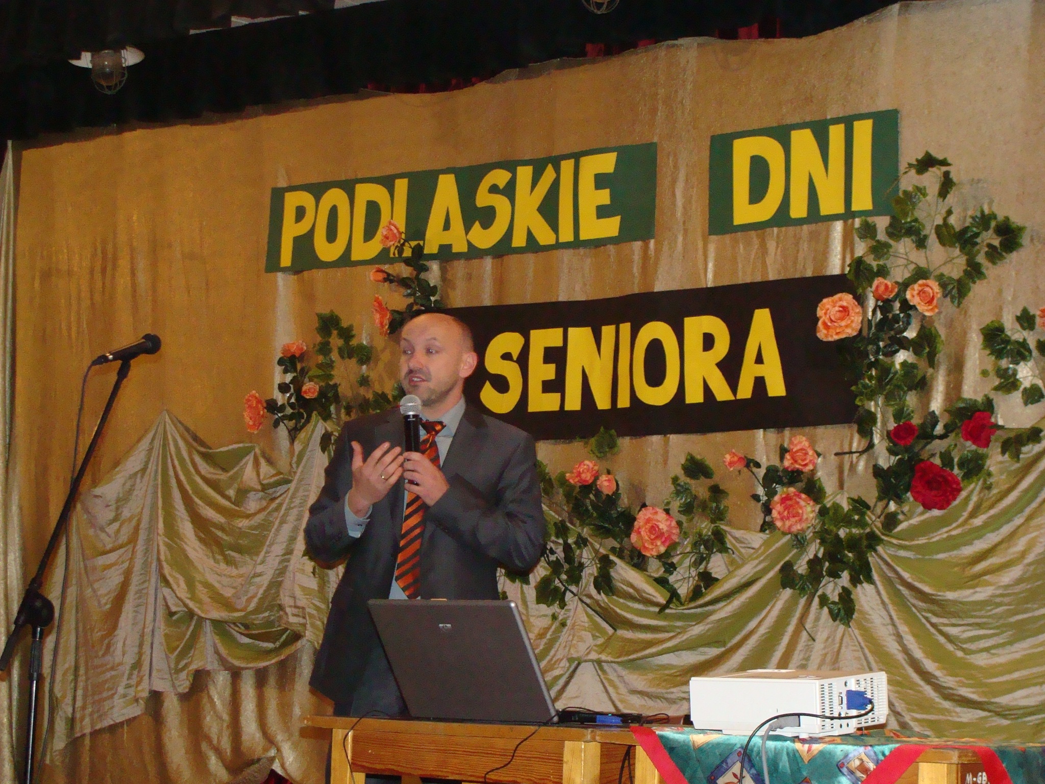 Mężczyzna stoi na scenie przy stole z laptopie i przemawia przez mikrofon.